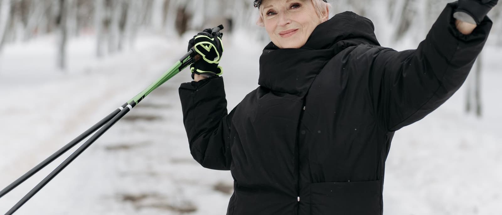 An older woman in winter clothing walking on a snowy path with poles, in bright flat daylight.