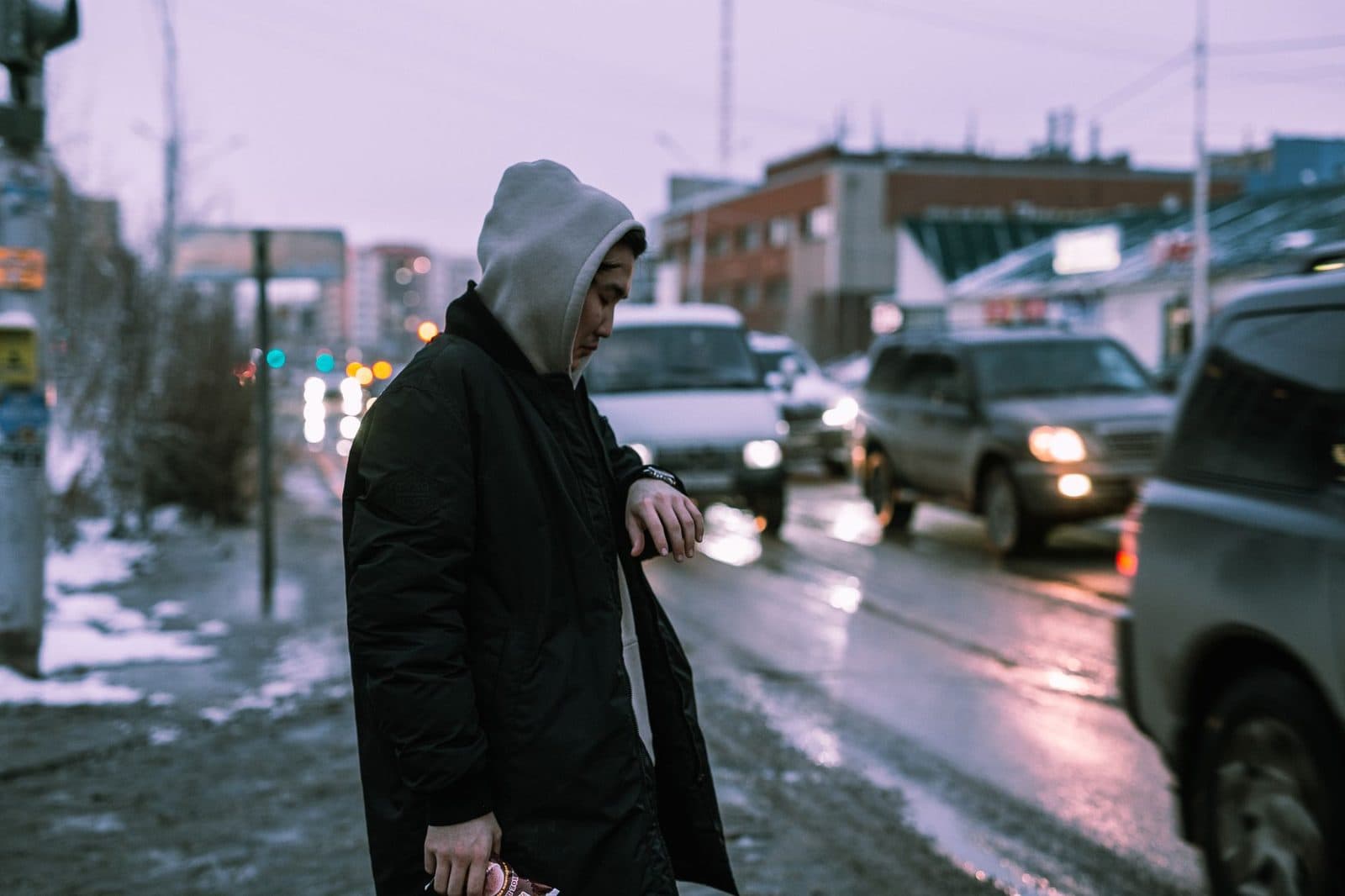 A person in a hoodie stands by roadside as traffic moves by on a chilly evening.