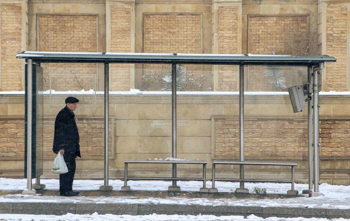 A person waiting at a bus stop on a gray winter morning, hands in pockets, pale flat light.