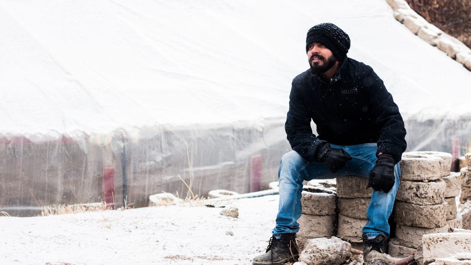 Man sitting on blocks in snowy outdoor setting, wearing winter clothing.