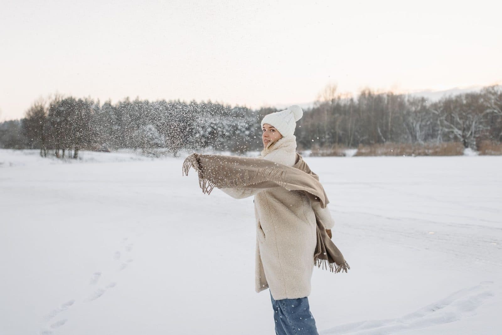A woman is playfully tossing snow in a serene winter landscape, dressed warmly in cozy clothing.