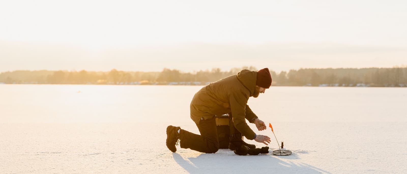 An angler kneeling beside a hole on a frozen lake in bright winter daylight.