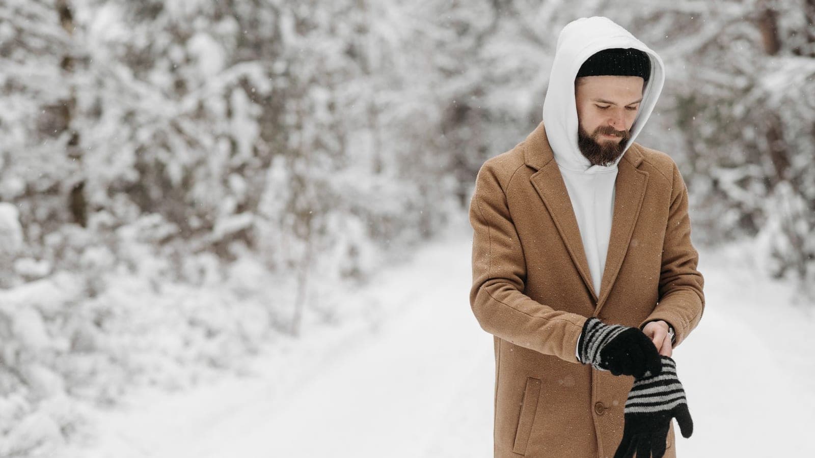 Bearded man in winter coat and gloves standing outdoors in snowy forest, surrounded by fresh snowfall.