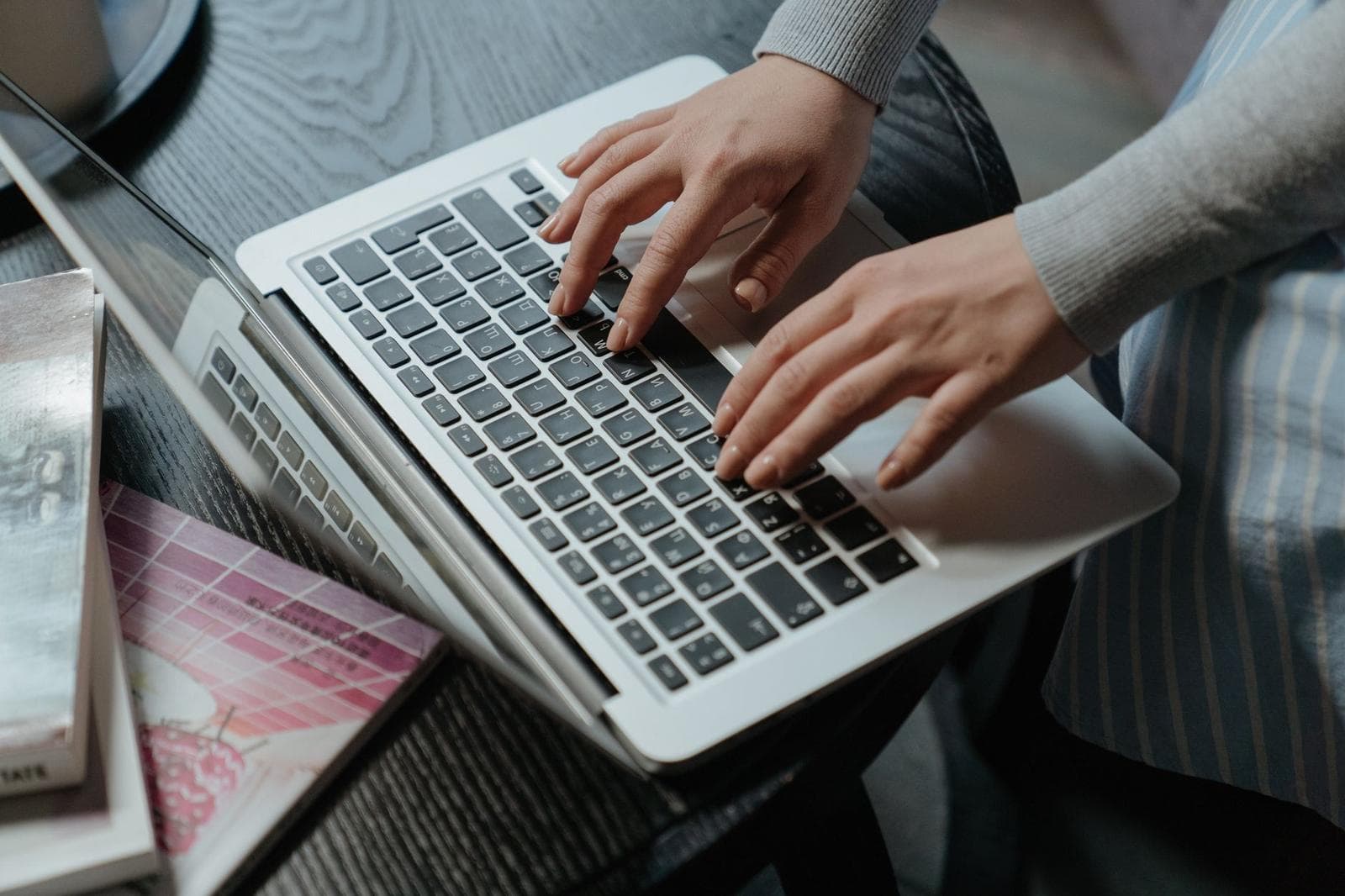 Close-up of a woman's hands typing on a laptop indoors, with books on the table.