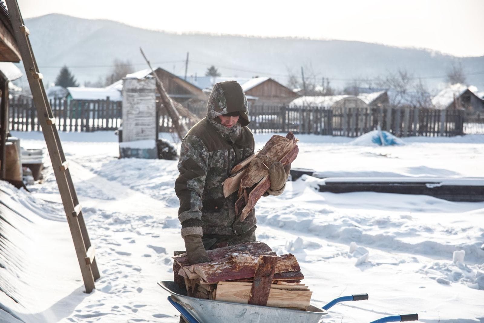 A man in winter clothing handles firewood in a snowy village landscape.