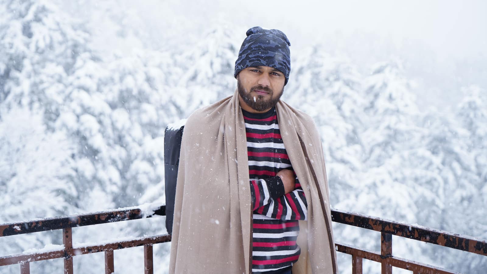 Man bundled in winter clothing standing amidst snowfall and a snowy forest backdrop.