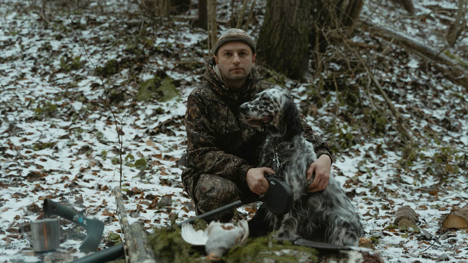 Man in camouflage with dog in a snowy forest, holding hunting equipment.