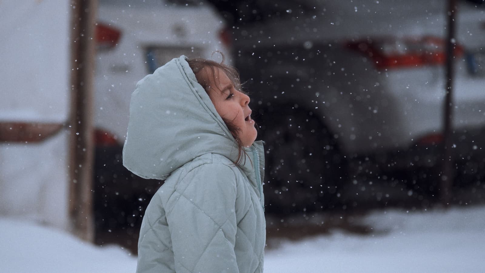 Young girl in a hooded coat marvels at falling snow during winter outdoors.