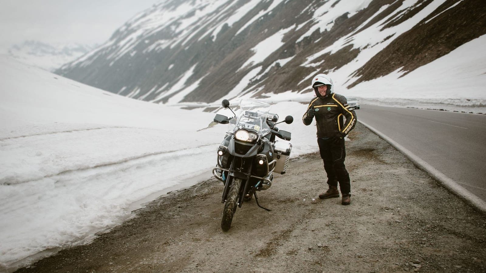 A motorcyclist stands beside his bike on a snowy mountain road, ready for adventure.