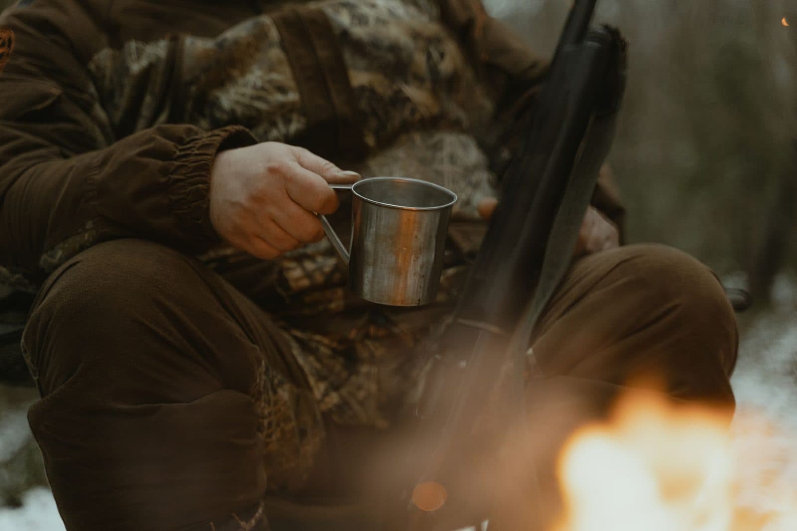A person in camouflage enjoys a campfire with a tin cup outdoors.