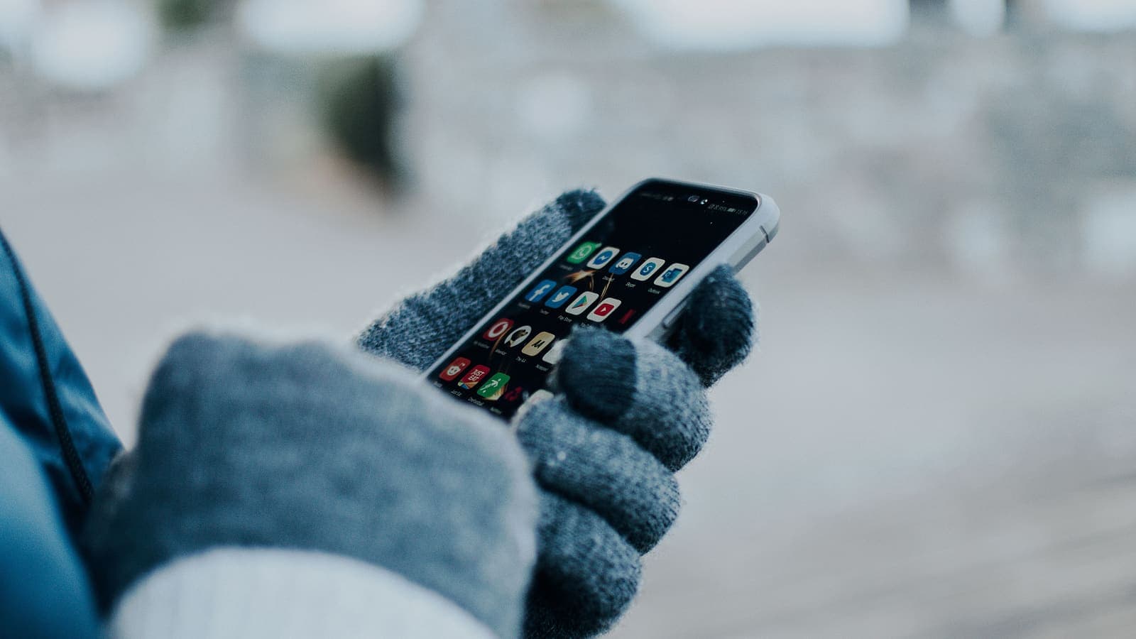 Close-up of hands using a smartphone with gloves in winter, showing apps on screen.