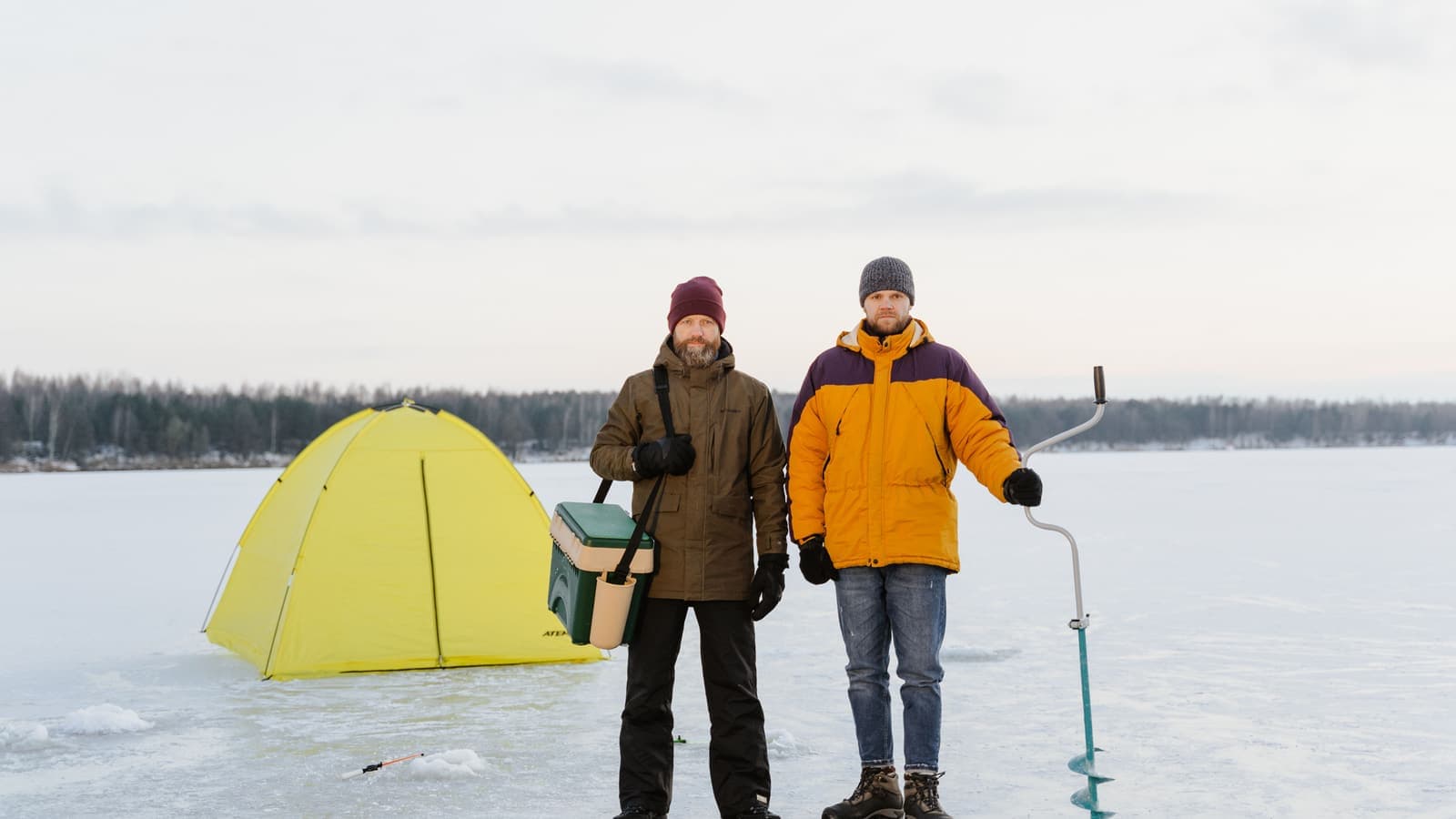 Two men ice fishing next to a tent on a frozen lake during winter.