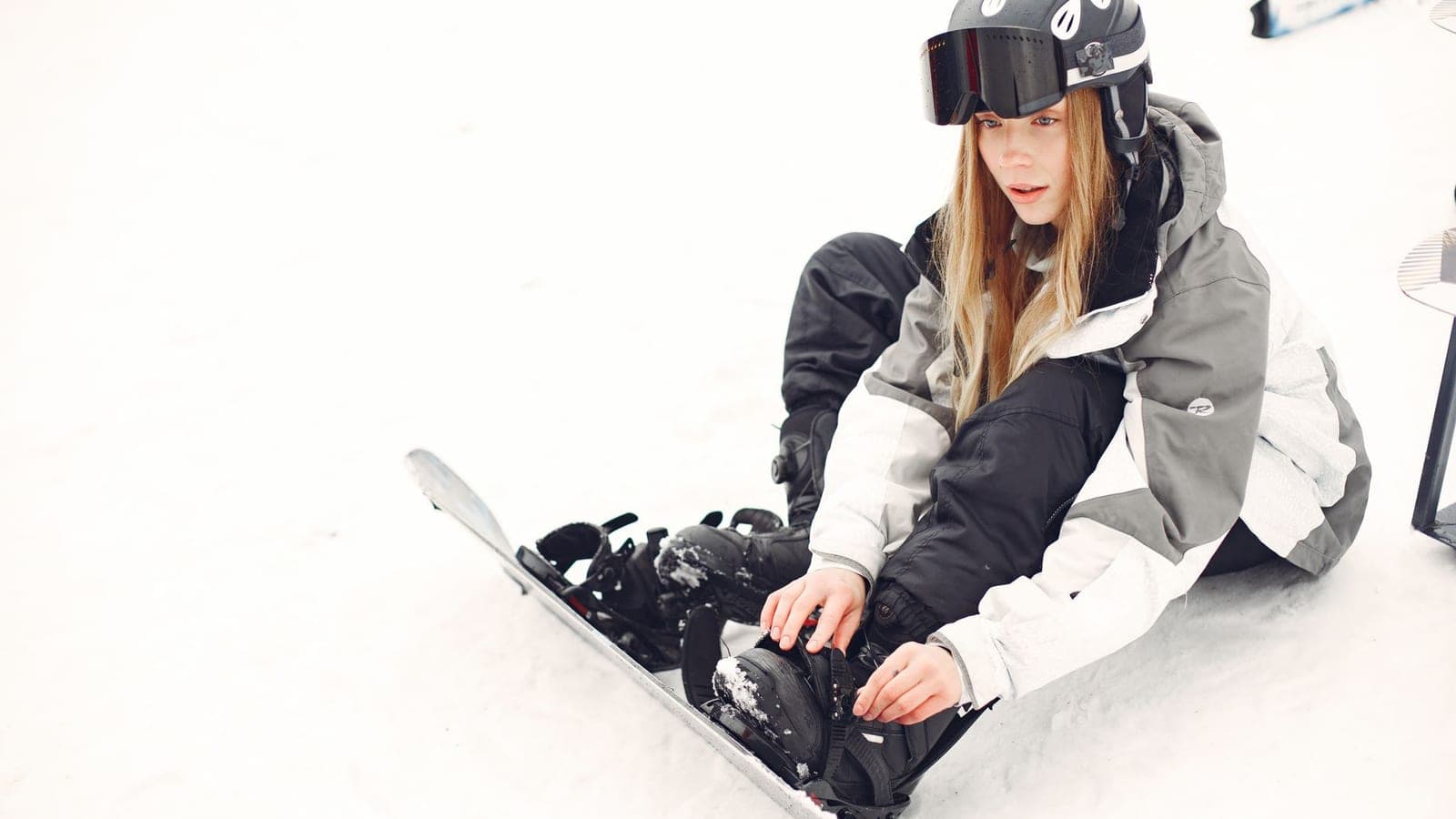A young woman snowboarder sitting on a snowy slope, adjusting her bindings on a winter day.