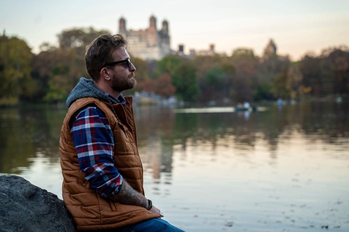 A man in a quilted vest over a flannel shirt on a cold overcast morning, pale sky and bare branches behind.